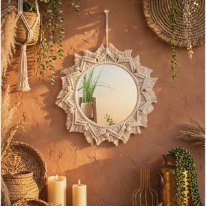 May include: A round wall mirror framed with off-white macrame cord featuring a sunburst pattern of intricate knots and fringe. The mirror is suspended by a matching macrame cord loop and reflects a green plant in a woven basket.