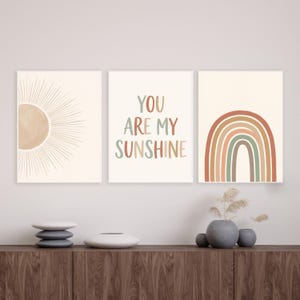 Pode incluir: Um conjunto de três impressões de parede com um sol com linhas radiantes, o texto YOU ARE MY SUNSHINE em letras multicoloridas e um arco-íris multicolorido. As impressões usam uma paleta de tons terracota, verde sálvia e creme.