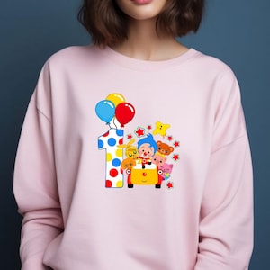 May include: A light pink sweatshirt featuring a colorful graphic. The graphic includes a large number one with polka dots, balloons, and cartoon characters in a yellow car. The balloons are blue, yellow, and red.