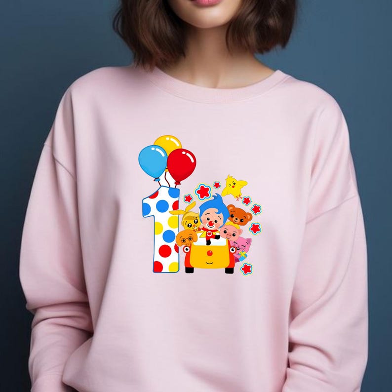 May include: A light pink sweatshirt featuring a colorful graphic. The graphic includes a large number one with polka dots, balloons, and cartoon characters in a yellow car. The balloons are blue, yellow, and red.