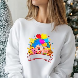 May include: White sweatshirt featuring a colorful cartoon graphic of a clown holding balloons, surrounded by other cartoon characters. The graphic includes a red banner at the bottom. The design is centered on the front of the sweatshirt.