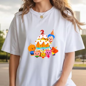 May include: White t-shirt featuring a colorful graphic of a birthday cake with the number 2 and cartoon characters. The cake is decorated with red cherries and yellow frosting. The characters include a dog, pig, rabbit, and bear.