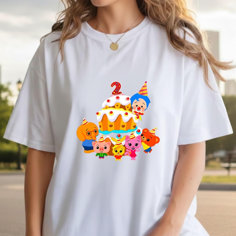 May include: White t-shirt featuring a colorful graphic of a birthday cake with the number 2 and cartoon characters. The cake is decorated with red cherries and yellow frosting. The characters include a dog, pig, rabbit, and bear.
