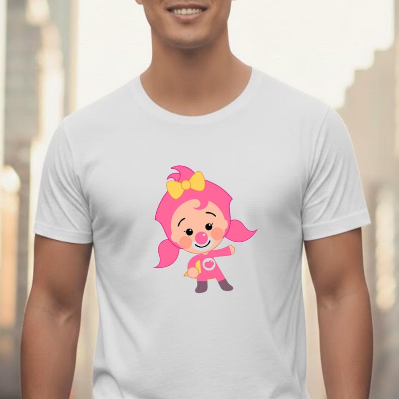 May include: White t-shirt featuring a cartoon character in a pink outfit with a yellow bow. The character has rosy cheeks and a smiling expression. The design is centered on the shirt.