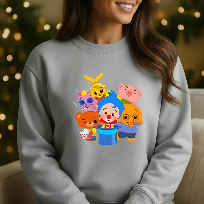 May include: A light gray sweatshirt featuring a colorful graphic of cartoon characters. The design includes a clown, a bear, a pig, an elephant, a cat, and a bunny. The characters are in bright colors, creating a playful and cheerful design.