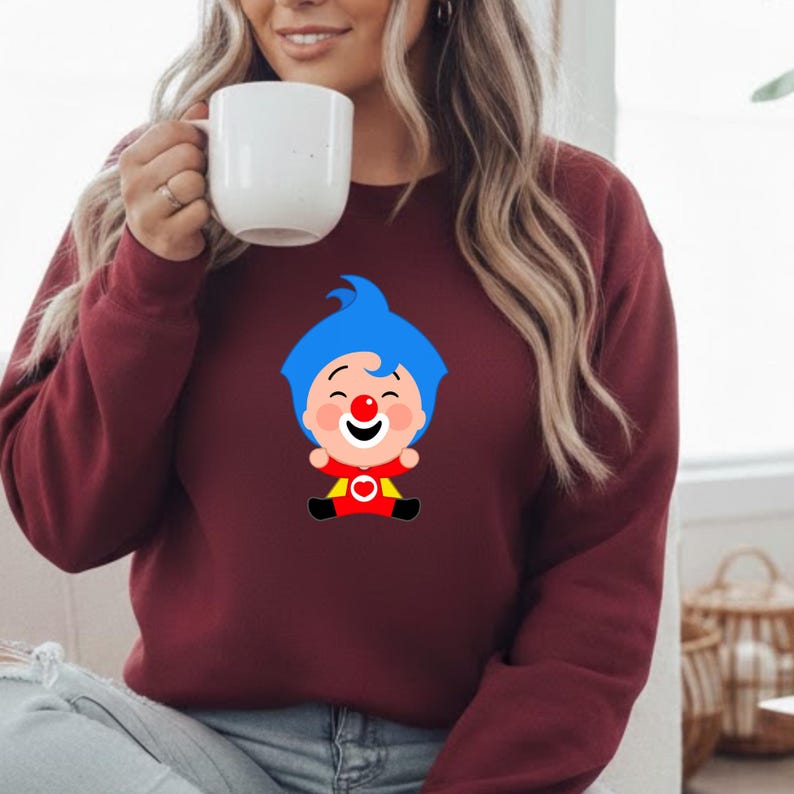 May include: Burgundy sweatshirt featuring a cartoon clown character with blue hair, a red nose, and a red and yellow outfit. The sweatshirt is a solid burgundy color and has a crew neck. The design is centered on the front of the shirt.