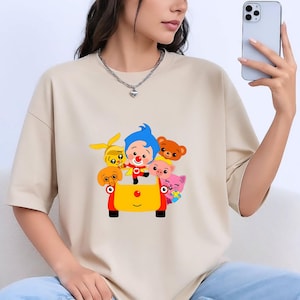 May include: Beige t-shirt featuring a colorful cartoon graphic of a clown and several animal characters in a yellow and red vehicle. The design includes a blue-haired clown, a yellow rabbit, a brown bear, a pink cat, and other cartoon characters.