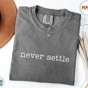 May include: A charcoal grey short-sleeve t-shirt with a crew neckline and the text "never settle" printed in white across the chest. The shirt features a Comfort Colors brand label at the inner neck and a faded, garment-dyed fabric finish.