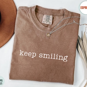 May include: An espresso-coloured short-sleeve t-shirt with a crew neckline. The shirt features the white text 'keep smiling' printed across the chest. A silver-toned necklace with a small circular pendant rests on the fabric near the collar.