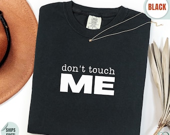 May include: A black crew neck t-shirt featuring the white printed text don't touch ME centered on the chest. The shirt has a Comfort Colors brand label at the inner neckline and is displayed flat with a thin gold chain necklace resting on the collar.