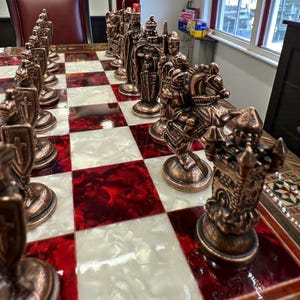 May include: A detailed chess set with bronze-colored pieces arranged on a red and white checkered board. The pieces include knights on horseback, castles, and other traditional chess figures. The board has a glossy finish.
