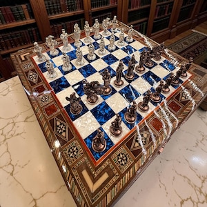 Könnte beinhalten: Ein Schachspiel mit einem Brett aus abwechselnd blauen und weißen perlmuttartigen Quadraten, umrandet von komplizierten Holzeinlegemustern. Das Set enthält metallisch silberne und bronzefarbene Schachfiguren mit detaillierten Skulpturen.