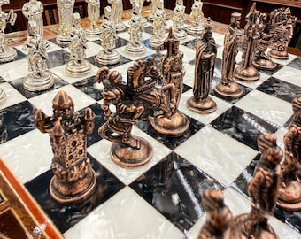 Op de afbeelding: Een schaakspel met metalen koperkleurige en zilverkleurige stukken op een schaakbord met afwisselend zwarte en witte vakken. De stukken zijn vormgegeven als gedetailleerde historische figuren, ridders te paard en torens.