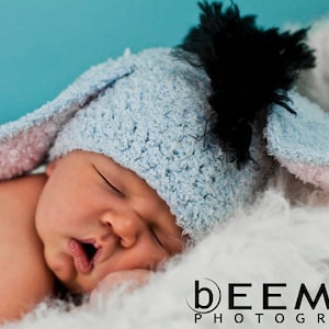 May include: A baby wearing a blue and pink crocheted bunny hat with black tulle accents. The baby is sleeping peacefully on a white fluffy surface.