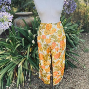 May include: High-waisted, wide-leg pants on a mannequin. The pants feature a floral pattern in shades of orange, yellow, and green on a cream background. The pants are displayed outdoors with greenery and flowers in the background.