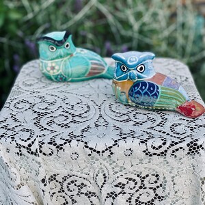 May include: Two hand-painted ceramic owls, one blue and one multi-colored, are sitting on a white lace tablecloth. The owls have detailed designs and are a decorative accent for a home.