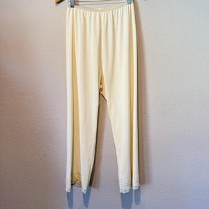 May include: A pair of cream-coloured lounge trousers featuring a smooth, solid fabric finish and an elasticated waistband. The bottom hem of each trouser leg is finished with a delicate, matching cream-coloured lace trim detail.