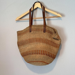 May include: A woven straw tote bag featuring a natural tan base with horizontal stripes in a burnt orange hue. The bag includes two dark brown leather handles securely attached with metal rivets, hanging from a wooden clothes hanger.