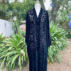 May include: A long, black velvet robe with a floral pattern is displayed on a mannequin. The robe has a V-neck and long sleeves. The background features greenery and a natural setting.
