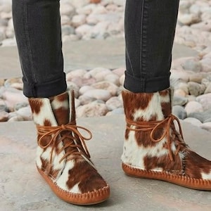 Può includere: Stivaletti mocassino alla caviglia con motivo a pelle di mucca marrone e bianco dalla consistenza pelosa. Gli stivali presentano una suola in cuoio color cuoio con cuciture a vista lungo il bordo e sottili lacci in pelle marrone annodati.