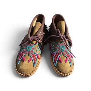 May include: A pair of tan suede moccasins featuring dark brown suede collars and fringe details. The vamps are decorated with intricate floral beadwork in vibrant shades of turquoise, magenta, and dark blue, finished with brown laces.