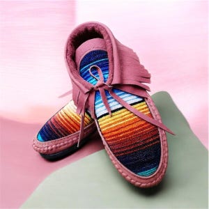 Pode incluir: Um par de mocassins malva com um padrão de riscas multicoloridas no peito do pé. Os sapatos têm franjas à volta do colarinho, um fecho de atacadores com tiras finas malva e um forro interior macio. O design incorpora faixas horizontais em azul, branco, amarelo, laranja e vermelho.