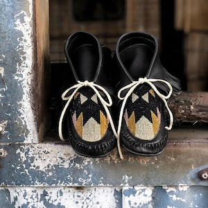 May include: A pair of black leather moccasins featuring fringe detailing and white laces. The top of each shoe is decorated with a geometric beaded pattern in black, gold, and cream colors. The moccasins have a visible whipstitch seam along the toe.