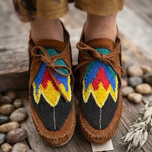 Op de afbeelding: Een paar bruine suède mocassins met een vetersluiting aan de voorzijde en een kleurrijk, met kralen bewerkt voorblad. Het kralenwerk toont een geometrisch patroon in zwart, geel, rood, blauw en groenblauw. De mocassins hebben een zichtbare naad en veters.