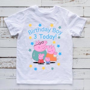 Op de afbeelding: Wit T-shirt met een cartoon varkensfamilie en de tekst "Birthday Boy 3 Today!" in het blauw. Het ontwerp bevat gele en blauwe sterren. Het shirt is gemaakt van een zacht, lichtgewicht materiaal.