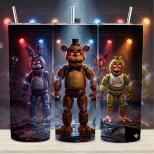 May include: A stainless steel tumbler featuring characters from the video game Five Nights at Freddy's. The design includes Bonnie, Freddy, and Chica, with a stage backdrop and lightning effects. The tumbler has a straw.