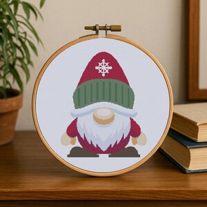 Puede incluir: Un bastidor de bordado de punto de cruz con un diseño de gnomo navideño. El gnomo lleva un gorro rojo con un copo de nieve blanco, un gorro de punto verde y una larga barba blanca. El cuerpo del gnomo es rojo, con manos y pies color canela.