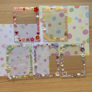 May include: Five clear, rectangular photo frames decorated with various embellishments. Each frame features a different design, including bows, hearts, stars, and pearls. The frames are arranged on patterned paper with stars and polka dots, creating a whimsical and decorative aesthetic.