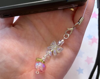 May include: A clear, pink apple charm with a green leaf and a silver clasp. The charm is attached to a silver chain with a flower and crystal bead accents. The charm is held in a hand.