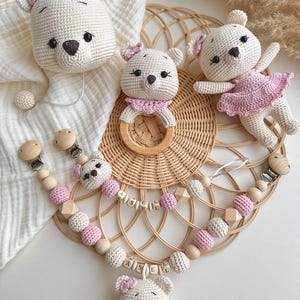 May include: A collection of handmade baby items in cream and pink. The image shows a crocheted bear mobile, a wooden ring rattle, and a pacifier clip with the name "ASEL" spelled out in wooden letters.