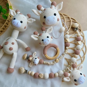 May include: A set of crochet giraffe toys and accessories including a stuffed giraffe, a rattle with a wooden ring, and two pacifier clips featuring wooden beads and the name MAEL JAMIL spelled out in white lettered cubes on the beaded strands.