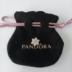 May include: A black velvet pouch with a pink drawstring, holding a silver flower-shaped charm. The charm is adorned with clear, sparkling stones. The word "PANDORA" is printed in gold on the pouch.
