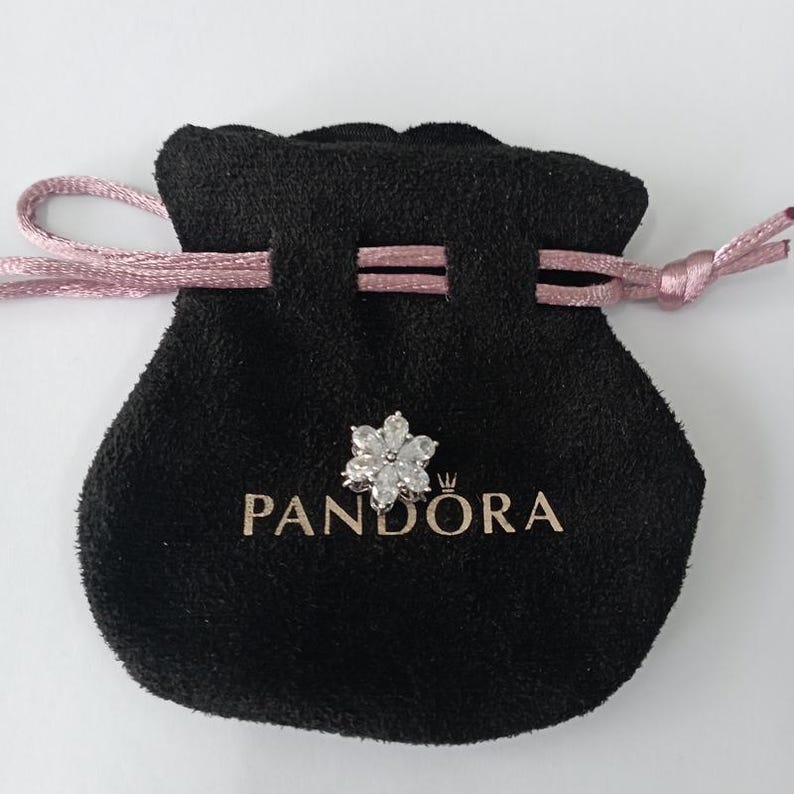 May include: A black velvet pouch with a pink drawstring, holding a silver flower-shaped charm. The charm is adorned with clear, sparkling stones. The word "PANDORA" is printed in gold on the pouch.