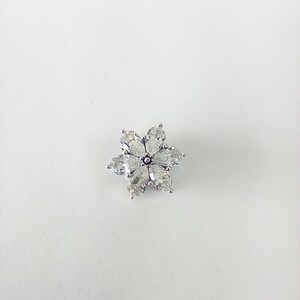 May include: A silver-toned flower-shaped brooch with six clear, teardrop-shaped crystals forming the petals. The center of the flower has a small, round, dark-colored accent. The brooch is set against a white background.