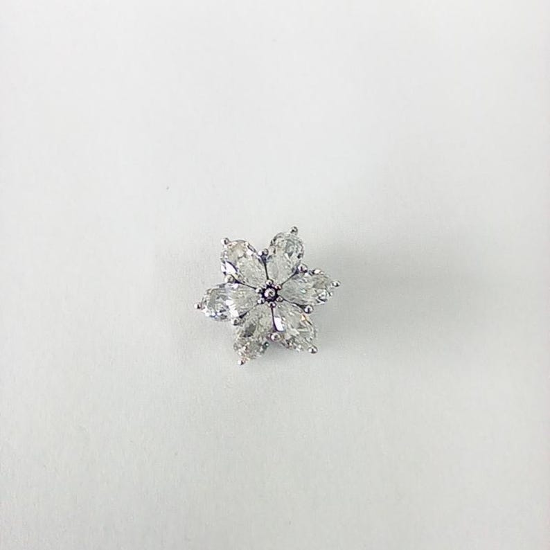 May include: A silver-toned flower-shaped brooch with six clear, teardrop-shaped crystals forming the petals. The center of the flower has a small, round, dark-colored accent. The brooch is set against a white background.