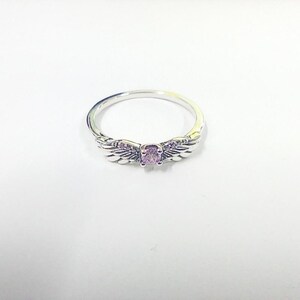 May include: A silver ring featuring a central, oval-shaped, light purple gemstone. The ring's design includes delicate wing motifs on either side of the gemstone, creating a celestial or angelic aesthetic. The ring is set against a white background.