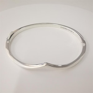 May include: A silver hinged bangle bracelet. The bracelet has a smooth, polished finish and a simple, elegant design. The clasp is visible on the left side. The bracelet is marked with "S 925 ALE PANDORA O".