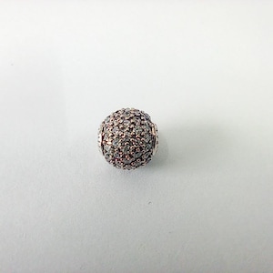Puede incluir: Una cuenta redonda de plata, cubierta de pequeñas piedras claras y brillantes. La cuenta tiene una superficie texturizada y está diseñada para la fabricación de joyas. La cuenta está sobre un fondo blanco.