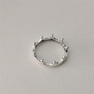 May include: A silver crown ring with a polished finish. The ring features a series of rounded points along the top, resembling a royal crown. The ring is set against a light gray background.