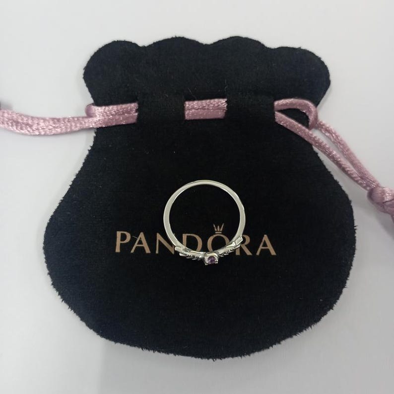 May include: A silver ring with a small purple gemstone, resting on a black velvet pouch. The pouch has a drawstring closure and the brand name "PANDORA" is printed on it. The ring is delicate and elegant.