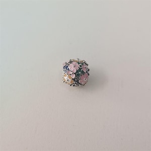 May include: A silver charm bead with a floral design. The bead is covered in small, colorful flowers in shades of pink, blue, green, and yellow. The flowers are arranged closely together, creating a textured surface.