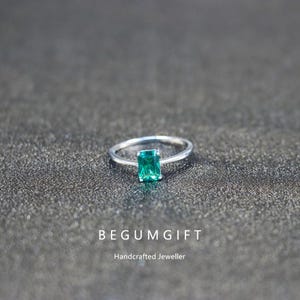 Peut inclure: Une bague en argent ornée d'une pierre précieuse rectangulaire vert émeraude. La bague est posée sur une surface sombre et texturée. Le texte "BEGUMGIFT" et "Handcrafted Jeweller" sont visibles en dessous.
