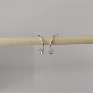 Op de afbeelding: Een paar zilverkleurige, gebogen oorbellen met kleine, heldere edelstenen aan de basis. De oorbellen worden getoond op een lichtgekleurde houten deuvel tegen een effen, lichtgrijze achtergrond. Het ontwerp is eenvoudig en elegant.