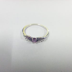 May include: A silver ring with a central oval-shaped, light purple gemstone. The ring features wing-shaped designs on either side of the gemstone, with small, embedded purple stones along the wings. The ring is set against a white background.