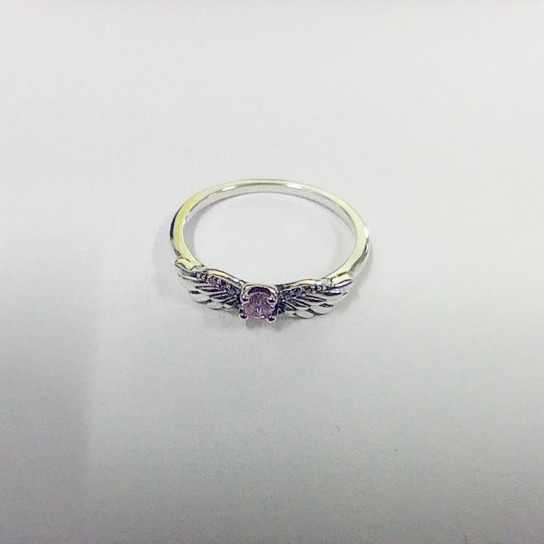 May include: A silver ring with a central oval-shaped, light purple gemstone. The ring features wing-shaped designs on either side of the gemstone, with small, embedded purple stones along the wings. The ring is set against a white background.