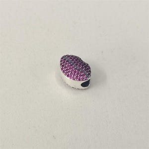 May include: A silver charm in the shape of a heart, encrusted with small, bright pink gemstones. A thin, dark blue line runs down the centre. The charm has a small, dark hole on one side, for a bracelet or necklace.